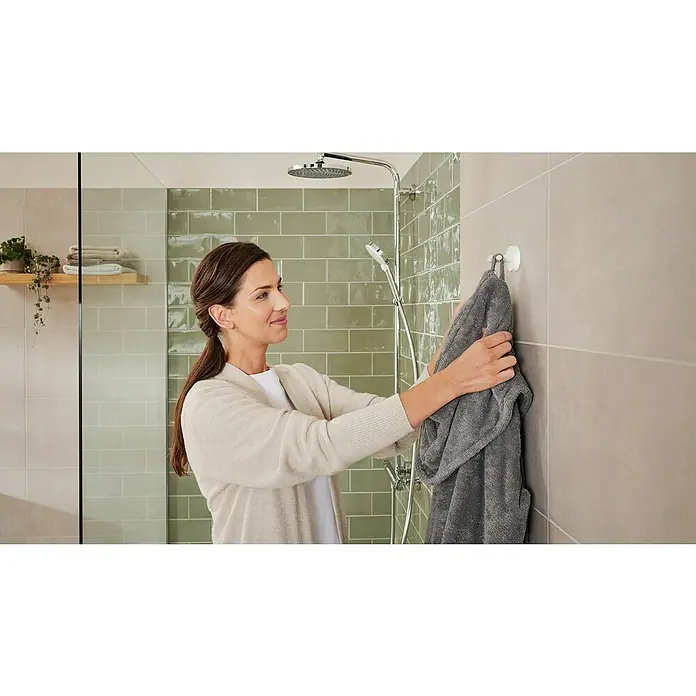 Tesa Moon Handtuchhaken - 2-armig, Weiß, Matt - Frau hängt ein graues Handtuch an einem Wandhaken im Badezimmer auf, mit einer Dusche im Hintergrund.