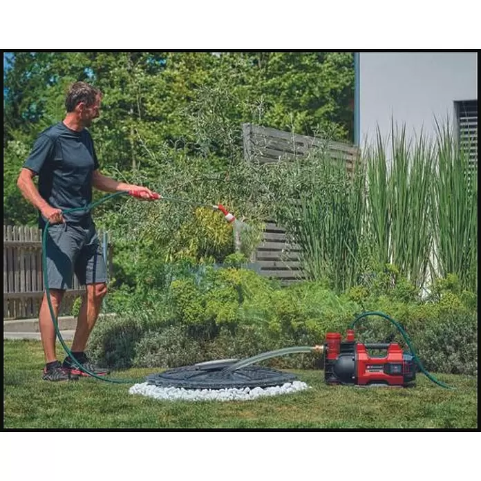Einhell PXC 18V Bomba de agua para jardín de batería 36/38 F LED Automatic - 18 V, Iones de litio, Sin batería, Capacidad de bombeo máx.: 3.800 l/h - Un hombre utiliza un sistema de bomba de jardín rojo y negro para regar plantas de un depósito subterráneo.
