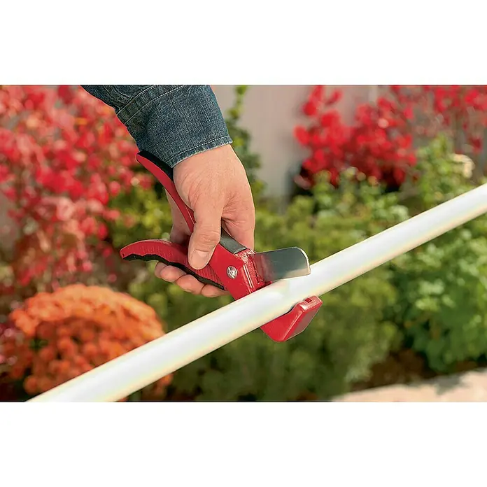 Cortatubos de plástico - Diámetro: 20 mm - 25 mm, Tubos de instalación - Primer plano de una mano usando un cortatubos rojo para cortar un tubo blanco, con plantas otoñales al fondo.