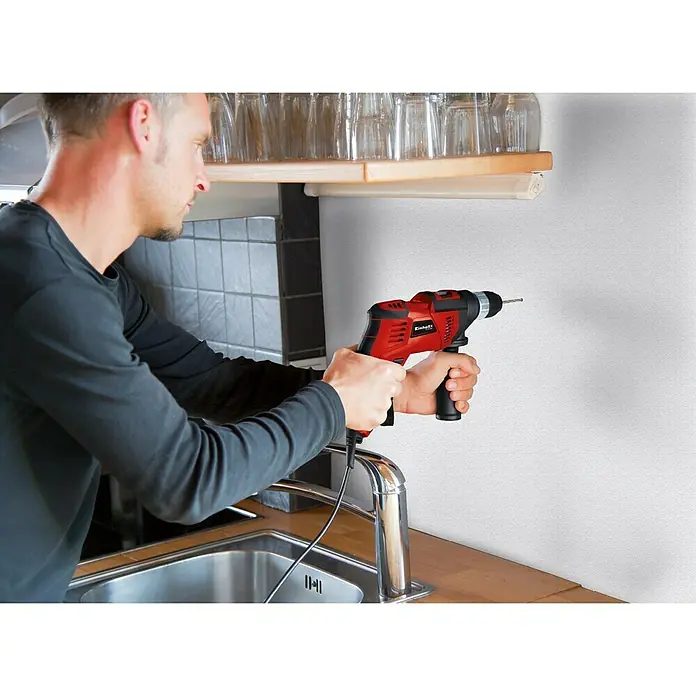 Einhell Schlagbohrmaschine TC-ID 1000 E Kit - 1.010 W, Schnellspannbohrfutter - Ein Mann bohrt mit einer roten Einhell Schlagbohrmaschine ein Loch in eine weiße Wand in einer Küche.