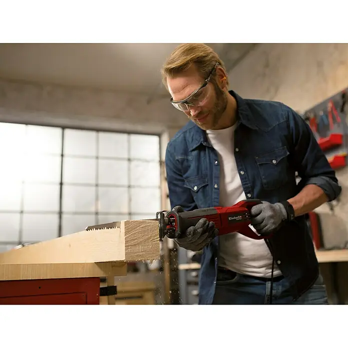 Einhell Universalsäge TE-AP 750 E - 750 W, Leerlaufhubzahl: 0 /min - 2.800 /min - Mann sägt Holz mit einer roten Einhell Säbelsäge, Schutzbrille und Handschuhen für Sicherheit.