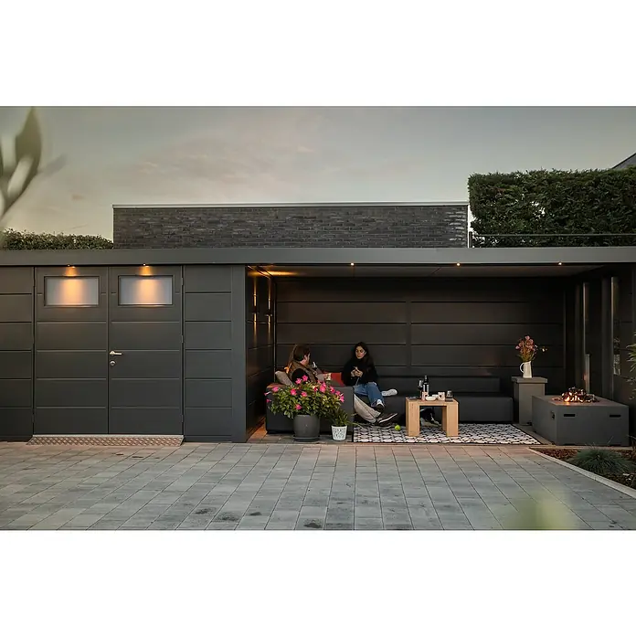 Telluria Metallgerätehaus Classico 3030 mit Lounge XL rechts inkl. 2 Fenster und Elektro-Set - Außenmaß inkl. Dachüberstand (B x T): 782 x 318 cm, Stahl, Anthrazit, Inkl. Innenwandverkleidung & Elektro-Set - Zwei Personen entspannen auf einer grauen Couch in einem stilvollen Außenbereich mit Beleuchtung und Pflanzen.
