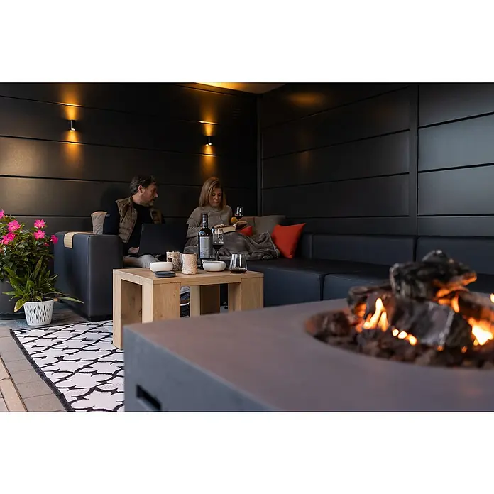Telluria Metallgerätehaus Classico 3330 mit Lounge XL rechts inkl. 2 Fenster und Elektro-Set - Außenmaß inkl. Dachüberstand (B x T): 812 x 318 cm, Stahl, Anthrazit, Inkl. Innenwandverkleidung & Elektro-Set - Entspannte Szene im Freien mit einer Feuerstelle im Vordergrund und einem Paar auf einem Sofa, das die gemütliche Atmosphäre genießt.