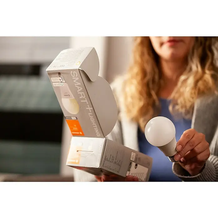 Ledvance Smart+ WiFi Smart-LED-Lampe - E27, Dimmbar, Mehrfarbig, 1.055 lm, 9,5 W - Eine Frau hält eine geöffnete Packung Smart+ Lighting und eine Smart+ Glühbirne in der Hand, die zum Auspacken bereit ist.