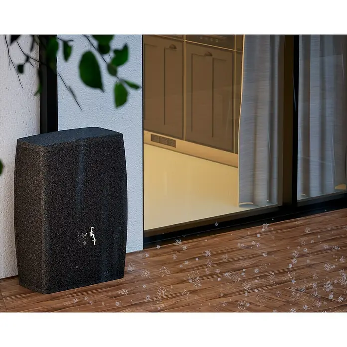 Arves Regenspeicher Boxy - 270 l, Granitkörnung, 1 Stk. - Ein stilvoller, dunkler Regenwassertank steht auf einer Holzterrasse vor einem Fenster, das einen Einblick in eine Küche bietet.