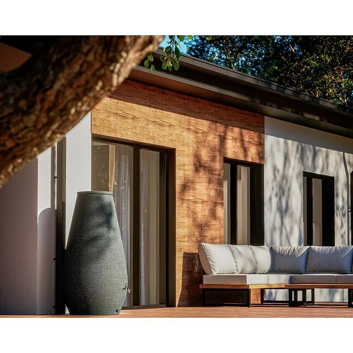 Arves Regenspeicher Drop - 410 l, Granitkörnung, 1 Stk. - Ein stilvoller Regenspeicher in Amphorenform neben einer modernen Hausfassade mit Sitzbereich. Ideal zur Gartenbewässerung und Wassersammlung.