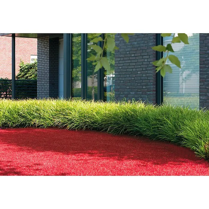 Rasenteppich Meterware Sunny - Breite: 200 cm, Rot, Mit Drainagenoppen - Das Bild zeigt einen Rasen mit rotem Kunstrasen vor einem modernen Haus mit grünen Büschen.