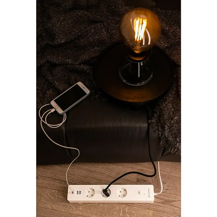Ledvance Smart+ WiFi Smart-Steckdosenleiste3-fach, Weiß, Max. Anschlussleistung: 3.680 W, 4 x USB - Das Bild zeigt eine weiße Leiste mit Steckdosen und USB-Anschlüssen, ein Smartphone, eine Lampe und einen Sofa.