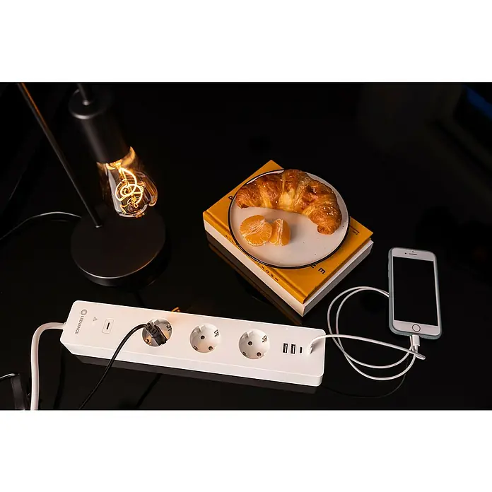 Ledvance Smart+ WiFi Smart-Steckdosenleiste3-fach, Weiß, Max. Anschlussleistung: 3.680 W, 4 x USB - Image of a LeDOUVANCE power strip with multiple outlets and USB ports, along with a croissant, tangerine, phone and lamp on a glossy black desk.
