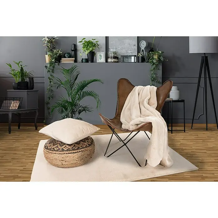 Decke Happy - Beige, 200 x 150 cm, 100 % Polyester - Einladendes Wohnzimmer mit einem Ledersessel, einer weichen Decke und einem Kissen auf einem Pouf, alles auf einem flauschigen Teppich.