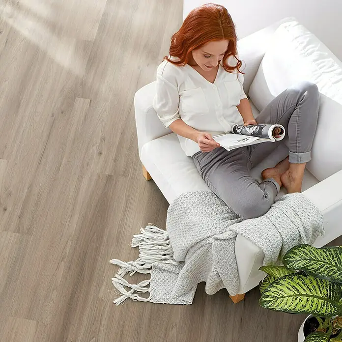 Decolife Designboden - Ash Oak, Landhausdiele, 1.220 x 185 x 10,5 mm - Frau entspannt sich auf einem Sofa mit einem Buch, wobei der Fokus auf dem hellen Holzboden liegt.