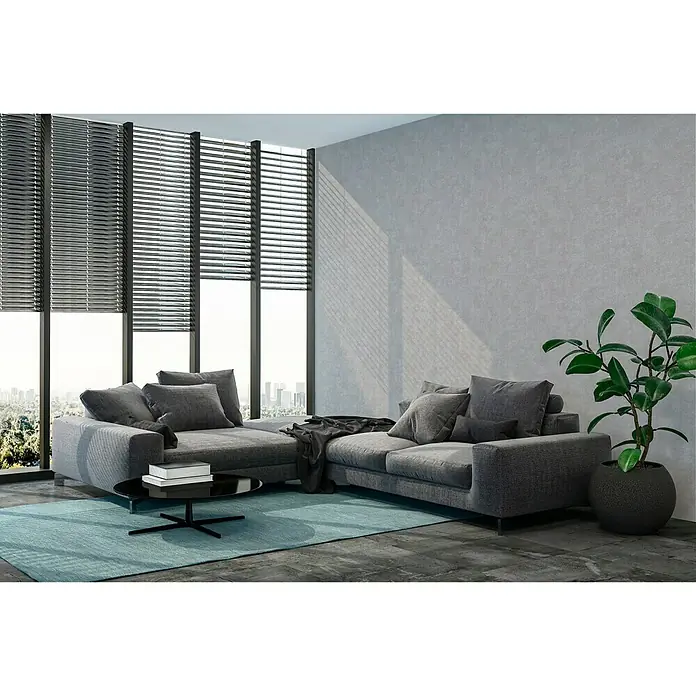 A.S. Création New Walls Vliestapete Beton - Grau, Betonoptik, 10,05 x 0,53 m - Ein modernes Wohnzimmer mit einem grauen Ecksofa, einem schwarzen Couchtisch und einem hellblauen Teppich vor einem großen Fenster.