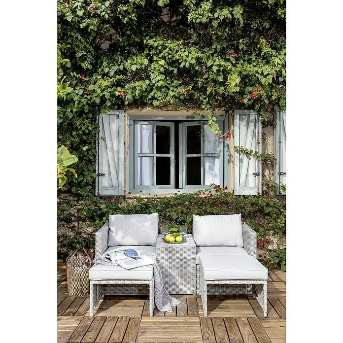 Conjunto de muebles Delma - 5 pzs., Ratán sintético, Gris - Acogedora escena al aire libre que muestra un juego de patio de mimbre blanco con dos sillones y otomanas, complementado con exuberante vegetación y flores.