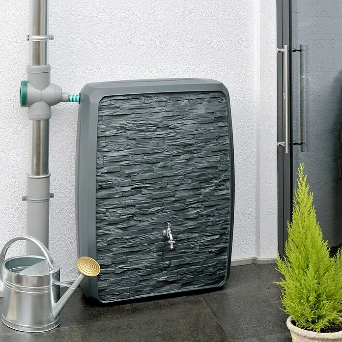 Arves Regenspeicher Multitank - 250 l, Anthrazit - Regenwasserbehälter in Steinoptik mit Wasserhahn, verbunden mit einem Fallrohr. Eine Gießkanne steht daneben, ein kleiner Baum im Topf.