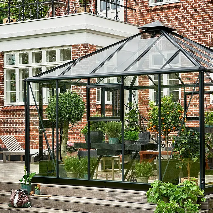 Juliana Gewächshaus Oase - Außenmaß inkl. Dachüberstand (B x T): 368 x 368 cm, Material Seitenwand: Glas, 3 mm, Anthrazit/Schwarz - Ein Gewächshaus mit Pflanzen und Bäumen auf einer Holzterrasse vor einem Backsteingebäude.