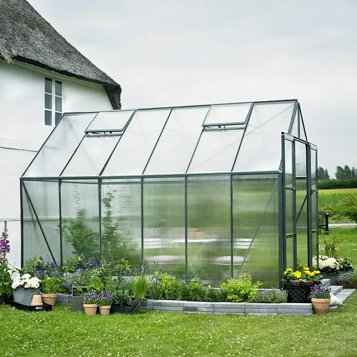 Halls Gewächshaus Magnum 128 - Außenmaß inkl. Dachüberstand (B x T): 257 x 384 cm, Material Seitenwand: Kunststoff, 6 mm, Grün - Ein geräumiges Gewächshaus mit transparenten Wänden und einem soliden Rahmen, umgeben von einem üppigen Garten mit Blumen und Pflanzen.