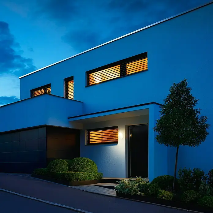 Steinel LED-Sensor-Strahler XLED home Slim S anthrazit - 7,2 W, L x B x H: 201 x 66 x 241 mm, Anthrazit, IP44 - Moderne Hausfassade in der Dämmerung mit beleuchteten Fenstern und gepflegtem Vorgarten, die architektonische Details hervorhebt.