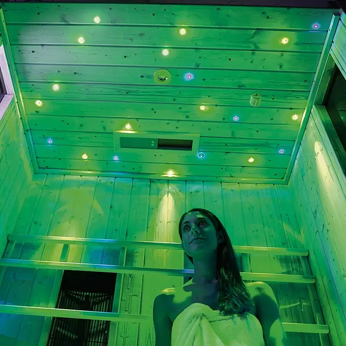 Weka Farbvisionen Set B - Anzahl LED: 43 Stk., Mindestraumhöhe: 240 cm - Eine Frau entspannt in einer Sauna mit LED-Beleuchtung, die eine beruhigende Atmosphäre schafft. Das Design der Sauna mit Holzdetails ist einladend.