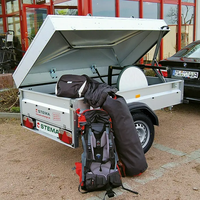 Stema Anhänger BASIC 750 mit Metalldeckel - Nutzlast: 575 kg, Einachser, Kasteninnenmaß: 201 x 108 x 47 cm, Ungebremst - Ein grauer Stema Anhänger mit offener Klappe und Gepäck, der für den Transport von Gegenständen verwendet wird.