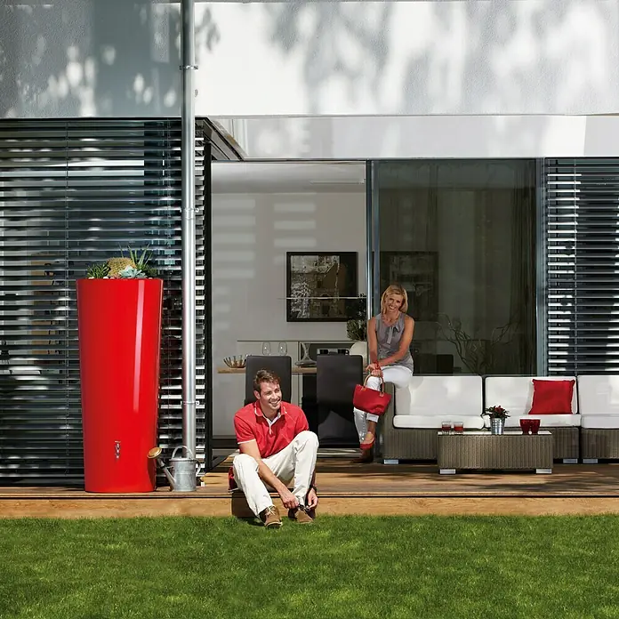 Garantia Modern Line Regenspeicher Color 2 in 1 - 350 l, Tomato - Stilvoller, roter Regenspeicher mit Pflanzen auf einer Terrasse, ergänzt durch eine silberne Gießkanne für den Garten.