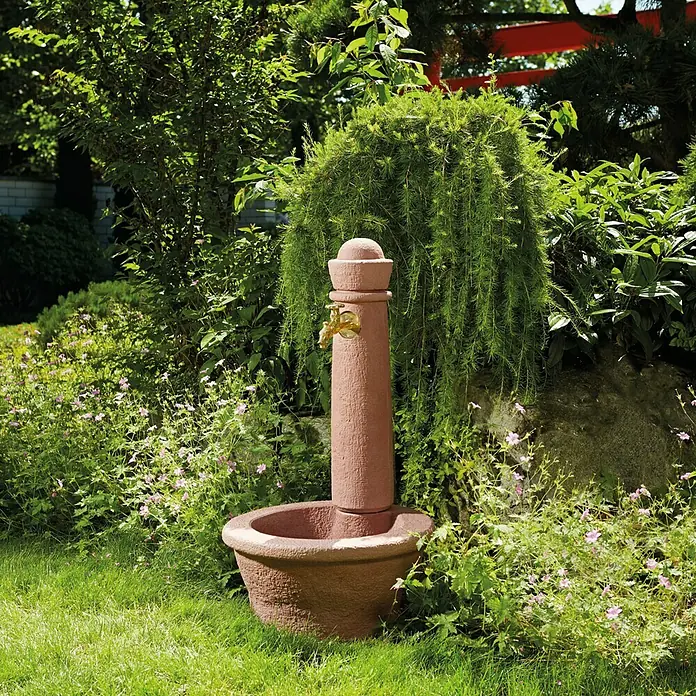 Garantia Brunnen Roma - Redstone, 54 x 54 x 100 cm - Rustikaler Gartenbrunnen aus Stein mit goldfarbenem Wasserhahn, umgeben von üppigem Grün.