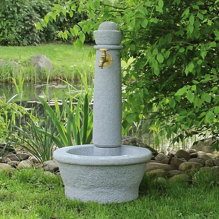 Garantia Brunnen Roma - Lightgranite, 54 x 54 x 100 cm - Ein dekorativer Steinbrunnen mit Messinghahn steht inmitten eines Gartens, umgeben von üppigem Grün. Ein Blickfang für jeden Garten.