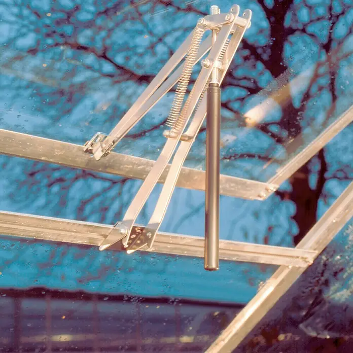 Juliana Fensteröffner Univent - Passend für: Juliana & Halls Gewächshäuser - Detailaufnahme eines automatischen Fensteröffners für Gewächshäuser, mit silbernen Metallarmen und einem schwarzen Zylinder vor einer verschwommenen Scheibe.