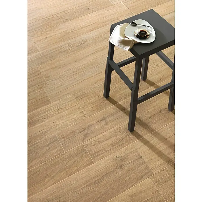 Harmonie by Palazzo Feinsteinzeugfliese - 30 x 120 cm, Eiche Hell, Matt - Das Bild zeigt eine Nahaufnahme von Keramikfliesen in Holzoptik, auf denen ein grauer Hocker mit einer Kaffeetasse steht. Die Fliesen verleihen dem Raum Wärme.