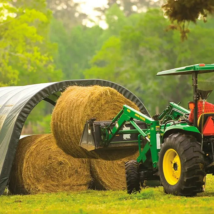 ShelterLogic Garage - Außenmaß inkl. Dachüberstand (B x T): 370 x 610 cm, Kunststoff, Grün - Ein grüner John Deere Traktor hebt einen großen Heuballen mit einem Frontlader vor einem Unterstand.