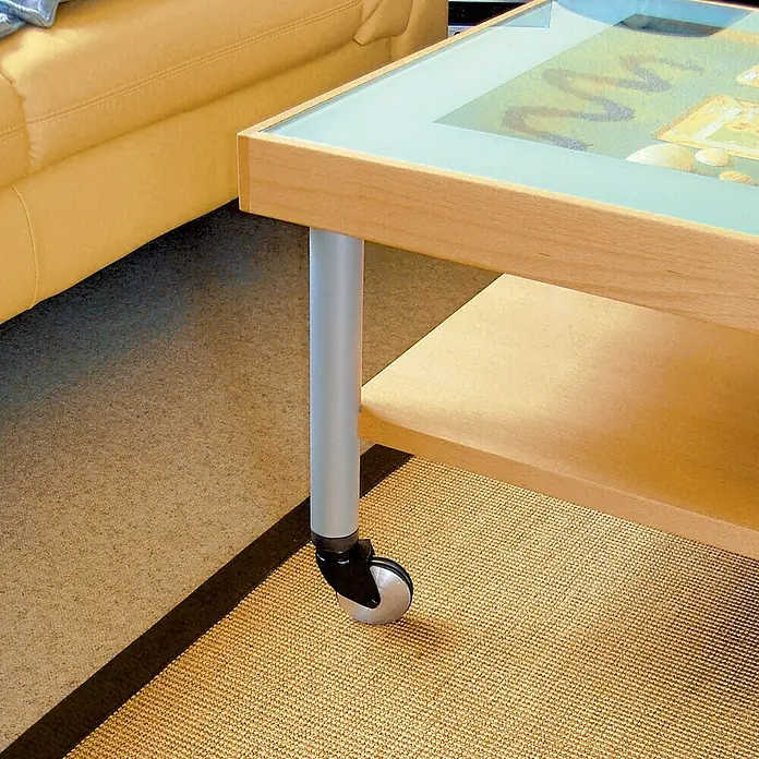 Dörner & Helmer Design-Lenkrolle - Durchmesser Rollen: 50 mm, Traglast: 50 kg, Gleitlager, Mit Platte - Nahaufnahme eines Couchtischs mit Glasplatte und Rollen, der neben einem gelben Sofa auf einem Teppich steht.
