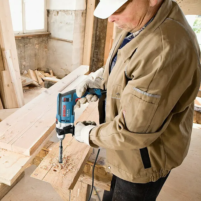 Bosch Professional Schlagbohrmaschine GSB 16 RE - 750 W, Schnellspannbohrfutter - Ein Mann bohrt mit einer blauen Bosch-Bohrmaschine ein Loch in ein Holzbrett. Gut geschützt mit Handschuhen und Arbeitskleidung.