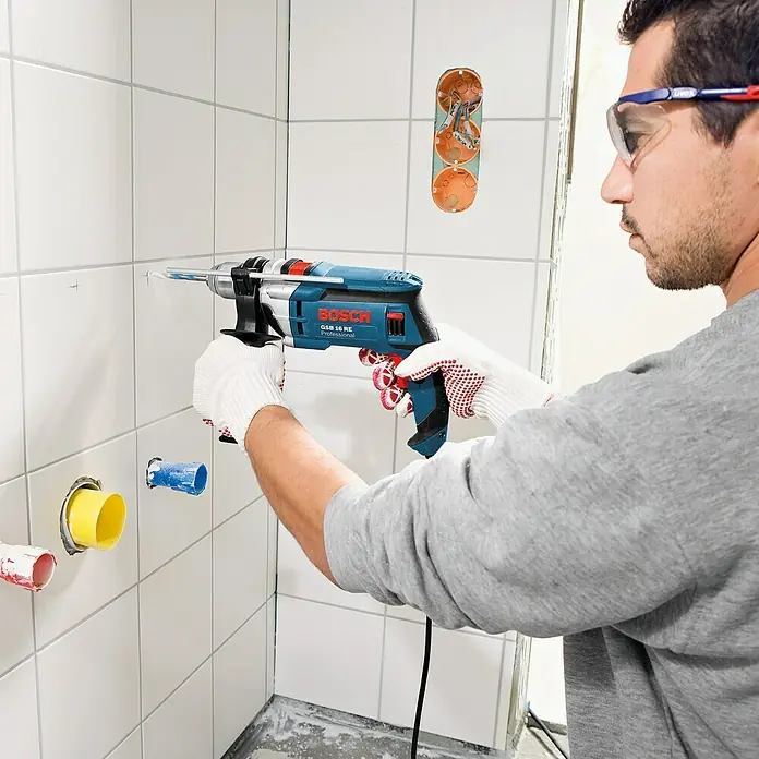 Bosch Professional Schlagbohrmaschine GSB 16 RE - 750 W, Schnellspannbohrfutter - Ein Mann bohrt mit einer Bosch GSB 16 RE Professional Bohrmaschine in eine geflieste Wand. Schutzbrille und Handschuhe werden getragen.