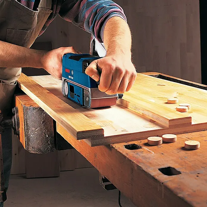 Bosch Professional Elektro-Bandschleifmaschine GBS 75 AE - 750 W, 200 m/min - 330 m/min - Der Bosch GBS 75 AE Bandschleifer wird zum Schleifen eines Holzstücks auf einer Werkbank verwendet, für präzise Holzarbeiten.