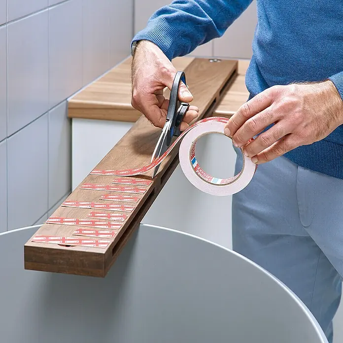 Tesa Montageband - Geeignet für: Fliesen, Belastbarkeit pro m: 100 kg, 1,5 m x 19 mm - Doppelseitiges Tesa-Klebeband wird mit einer Schere auf einem Holzbrett, das über einem Mülleimer liegt, zugeschnitten.