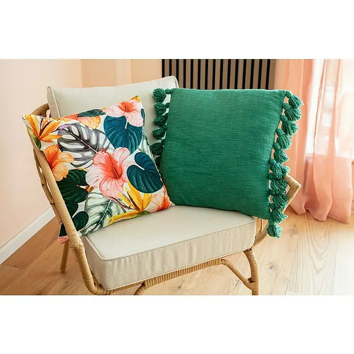 Kissen Arcos - Bunt, 45 x 45 cm, 100 % Baumwolle - Dekorative Kissen auf einem Rattansessel, darunter ein Kissen mit tropischen Blumen und ein grünes Kissen mit Quasten.