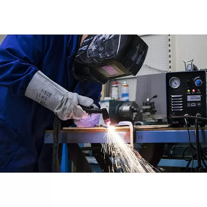 Güde Schweißhandschuh 6120 - Grau, Leder, L x B x H: 165 x 20 x 365 mm - Arbeiter in blauer Arbeitskleidung, Schweißhelm und Handschuhen benutzt einen Plasmaschneider, der helle Funken erzeugt.