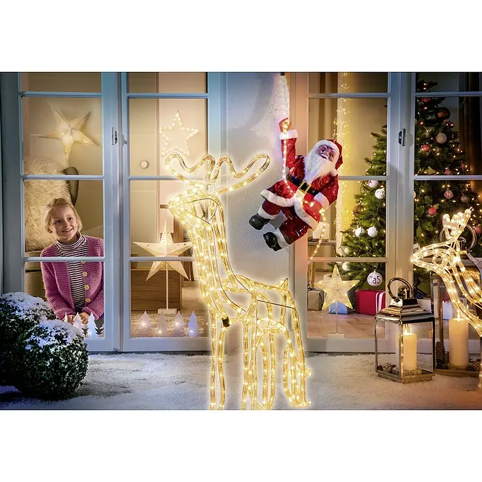 Tween Light Árbol de Navidad de LEDs Árbol de Navidad de papel - Para interior, 12 ud., Color de luz: Blanco cálido, Funciona con pilas - Escena festiva con renos iluminados, un Papá Noel en una escalera y una niña mirando por la ventana.