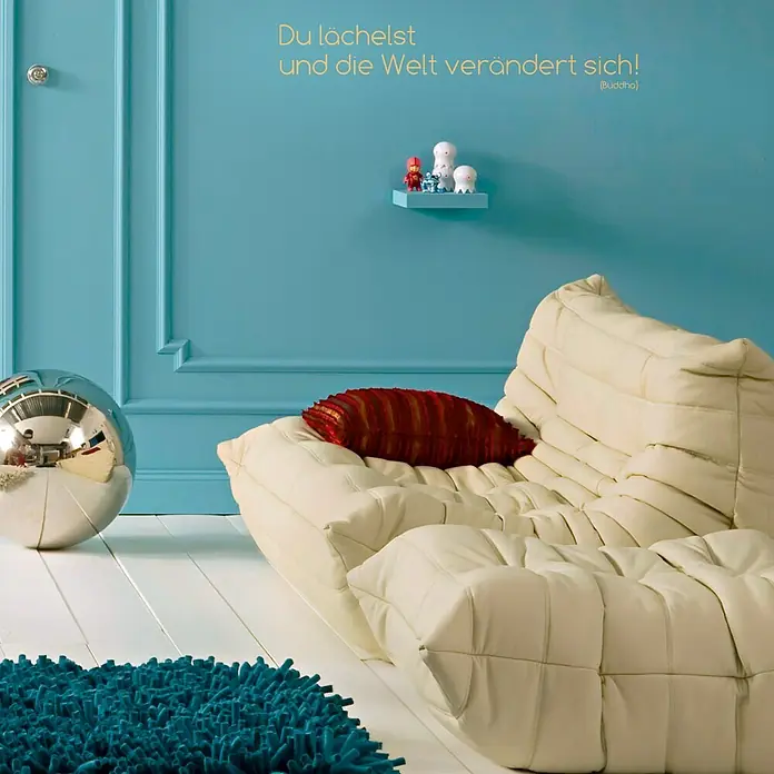 Komar Dekosticker Du Lächelst - 3 -tlg., 70 x 14 cm - Elegantes Interieur mit beigem Ledersofa, rotem Kissen, blauem Teppich und silberner Kugel. Zitat an der Wand rundet das Design ab.