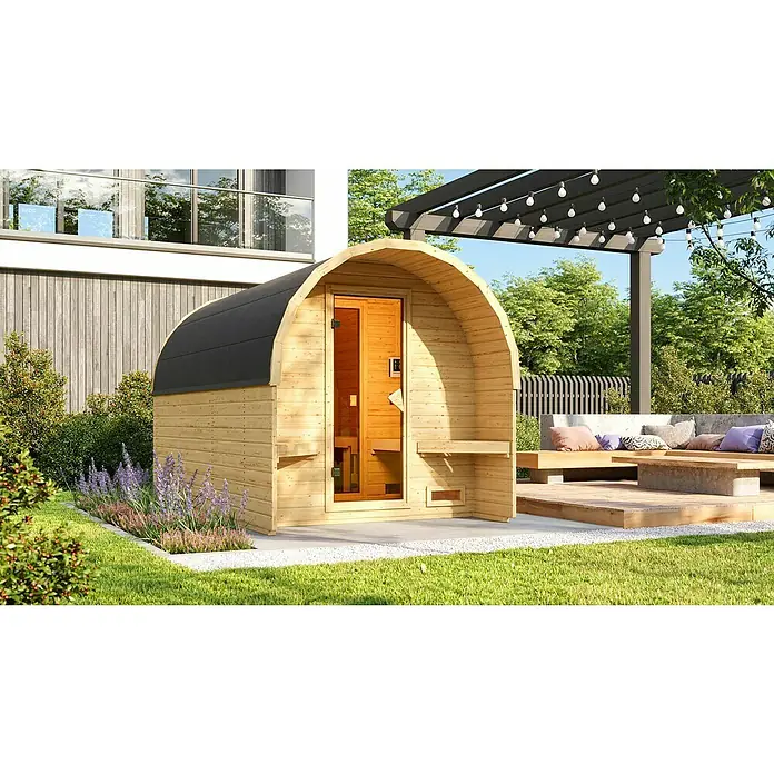 Karibu Fasssauna - L x B x H: 385 x 219 x 229 cm, Ohne Ofen - Ein freistehende Fasssauna aus Holz mit schwarzem Dach, umgeben von Grün und Blumen, lädt zur Entspannung im Garten ein.