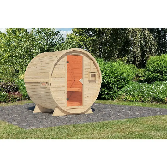 Karibu Fasssauna - L x B x H: 175 x 225 x 229 cm, Ohne Ofen, Anzahl Liegen: 2 Stk. - Eine runde Fasssauna aus hellem Holz mit Tür und kleinem Fenster steht im Garten auf einem gepflasterten Bereich. Die Sauna lädt zur Entspannung ein.