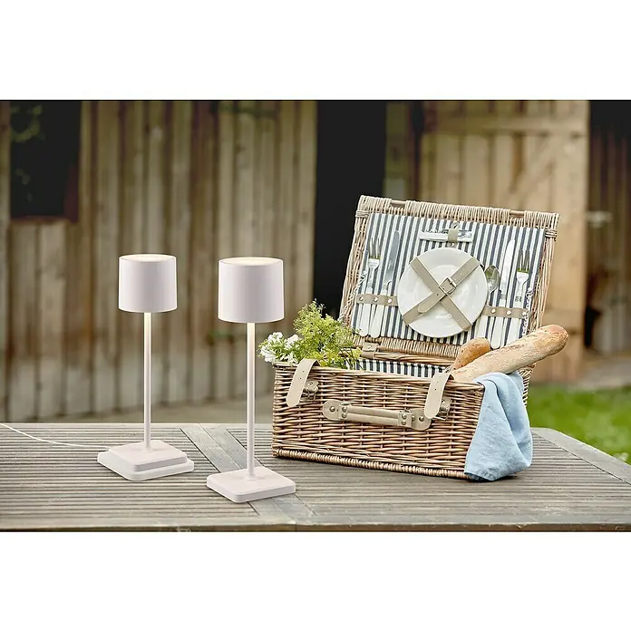 Reality Leuchten Akku-Tischleuchte FERNANDEZ - 1 W, L x B x H: 12,5 x 12,5 x 38 cm, Weiß, Mehrfarbig - Zwei kabellose Tischlampen neben einem Picknickkorb auf einem Holztisch im Freien. Die Lampen strahlen ein sanftes Licht aus.