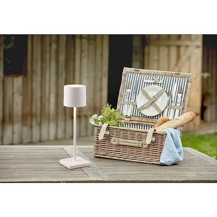 Reality Leuchten Akku-Tischleuchte FERNANDEZ - 1 W, L x B x H: 12,5 x 12,5 x 38 cm, Grau, Mehrfarbig - Eine schnurlose Tischleuchte mit einem Picknickkorb auf einem Holztisch im Freien, die eine gemütliche Atmosphäre für ein Picknick schafft.