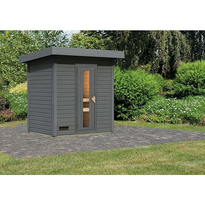 Karibu Saunahaus Hytti - Ohne Ofen, Terragrau, L x B x H: 146 x 196 x 228 cm - Eine graue Gartensauna mit Flachdach und Glastür auf einem gepflasterten Bereich, umgeben von Grün. Ideal für Entspannung im eigenen Garten.