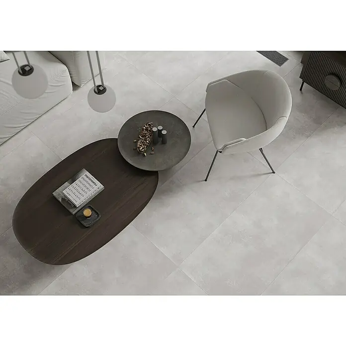 Feinsteinzeugfliese Big Show XXL - 59,8 x 119,8 cm, Grau, Matt - Eine moderne Sitzecke mit einem hellgrauen Sessel, einem dunklen, ovalen Couchtisch und einem runden Beistelltisch auf einem hellgrauen Fliesenboden.