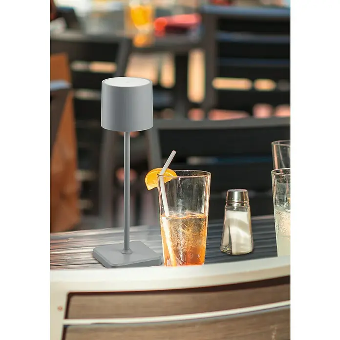 Reality Leuchten Akku-Tischleuchte FERNANDEZ - 1 W, L x B x H: 12,5 x 12,5 x 38 cm, Grau, Mehrfarbig - Eine graue kabellose Tischleuchte steht auf einem Tisch neben Getränken, die eine entspannte Atmosphäre erzeugen.