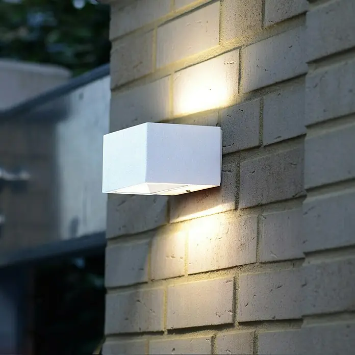 Lutec Aplique exterior LED Gemini - 10,5 W, 14 x 9 x 6,5 cm, Blanco, IP54 - Moderna lámpara de pared exterior blanca en una pared de ladrillos, emitiendo una luz cálida y acogedora. Diseño simple y elegante.