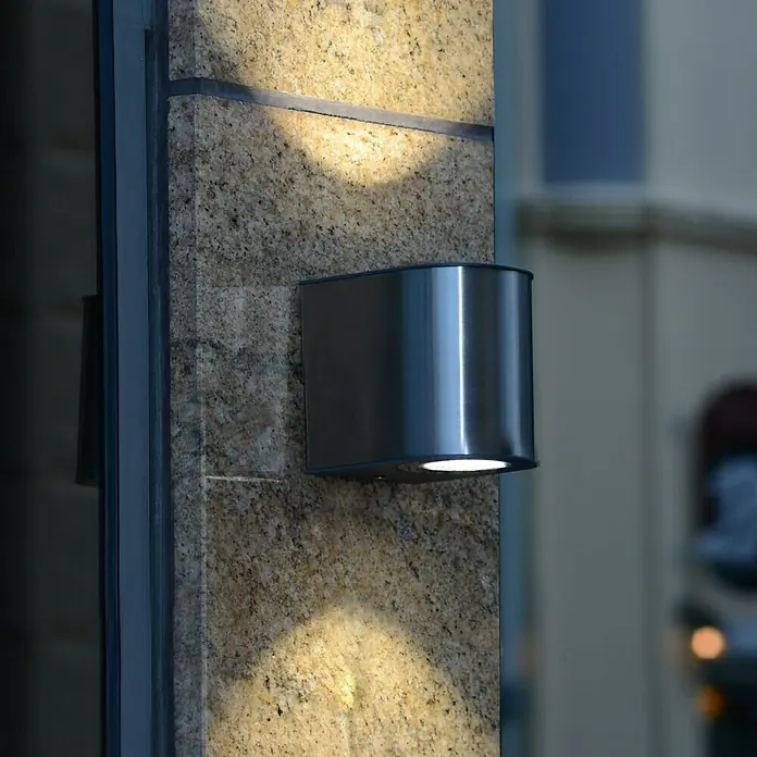 Lutec Aplique exterior LED Gemini - 9 W, 8 x 6,8 x 9,7 cm, Acero inoxidable, IP54 - Primer plano de un moderno aplique de pared exterior de metal que emite un brillo cálido sobre una pared de piedra.