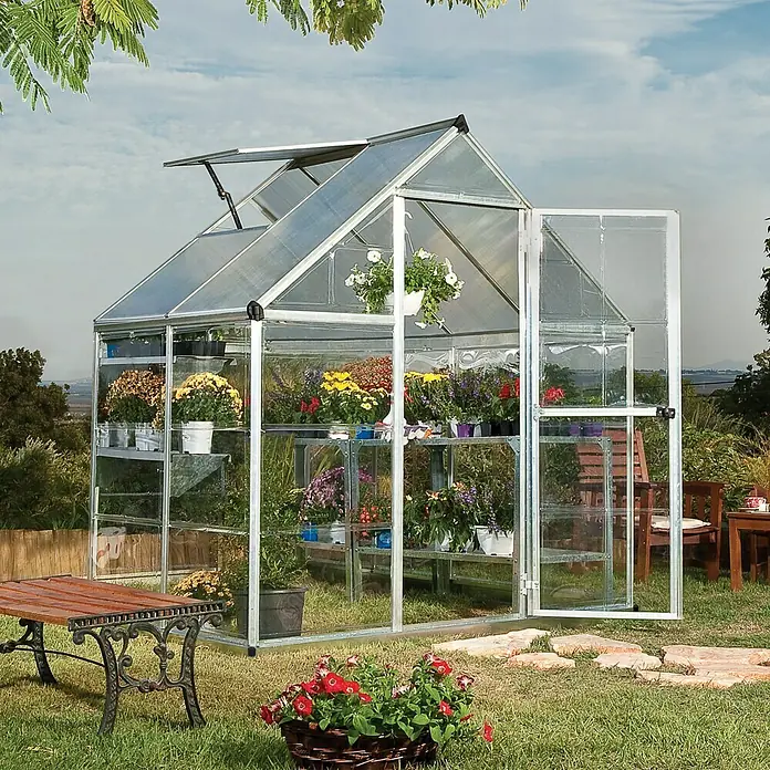 Palram – Canopia Gewächshaus Hybrid - Außenmaß inkl. Dachüberstand (B x T): 185 x 126 cm, 0,7 mm, Silber - Ein Gewächshaus aus Aluminium und Glas steht im Garten, gefüllt mit bunten Blumen auf Regalen. Die Tür ist geöffnet.
