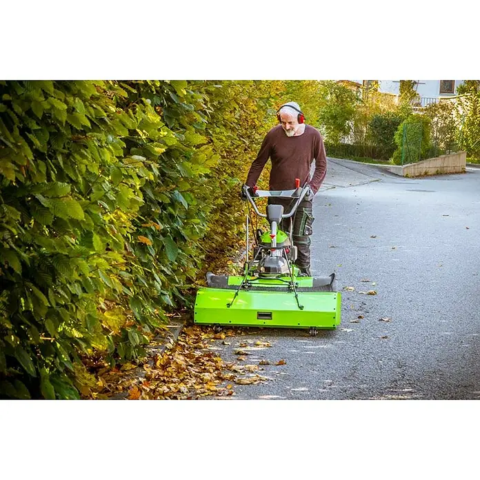 Zipper Kehrmaschine ZI-KM1000 - 3.600 W - Ein Mann benutzt eine grüne Kehrmaschine auf einer Straße, um Blätter zu entfernen, während er Kopfhörer trägt. Praktisches Gerät für die Gartenarbeit!