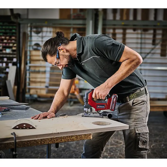 Einhell Professional Power X-Change 18V Akku-Pendelhubstichsäge TP-JS 18/135 Li BL - Solo - 18 V, Anzahl Akkus: Ohne Akku, 3.500 /min - Ein Mann benutzt eine Einhell Akku-Stichsäge, um ein Loch in ein Holzbrett zu schneiden. Er trägt dabei Schutzbrille und Gehörschutz.